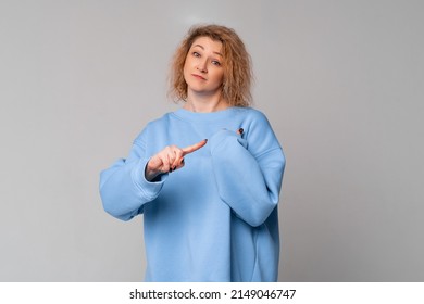 Serious middle age woman with curly blonde hair shakes her finger threateningly, scolding or telling off someone, standing in trendy blue sweatshirt over light grey background