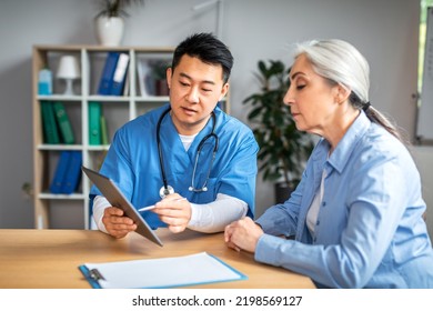 Serious Middle Aged Asian Man Doctor Showing Tablet To Retired European Woman Patient In Clinic Office Interior. Life Insurance, Treatment With Therapist, Medical Health Care, Receipt And Service