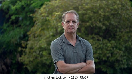 Serious Middle Aged Man Standing At Park Looking At Camera. Portrait Of An Older Male Person With Arms Crossed