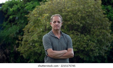 Serious Middle Aged Man Standing At Park Looking At Camera. Portrait Of An Older Male Person With Arms Crossed