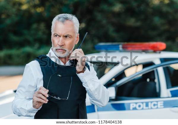 serious middle aged policeman in\
bulletproof vest talking on radio set near car at\
street