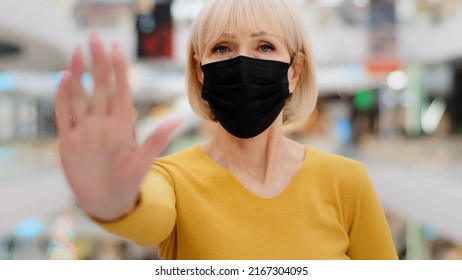 Serious middle-aged caucasian woman in medical mask protects health holding hand in front put palm forward showing stop gesture looking at camera keep safe distance during pandemic forbids approaching