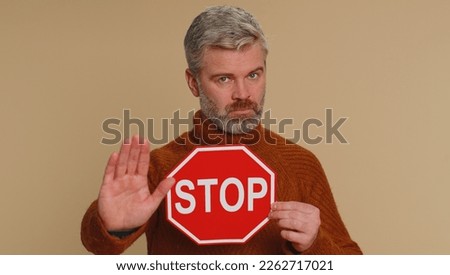 Serious middle-aged man say No, hold inscription text red No stop danger sign, warning of finish, prohibited access declining communication, body language, trouble, protest. Guy on beige background