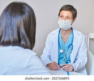 Serious Middle-aged Woman General Practitioner GP In Face Mask Consulting Female Patient During Covid-19 Outbreak, Doctor Giving Recommendations And Explaining Medical Diagnosis To Client In Clinic