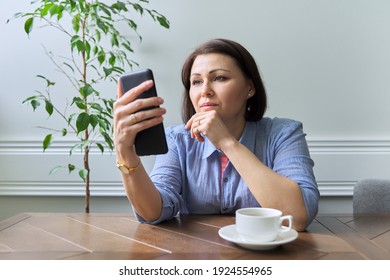 Serious Middle-aged Woman With Smartphone Sitting At The Table At Home. Mature Woman Looking At Phone Screen With Concentration, Video, Chat, Social Network, Important Information