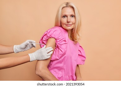 Serious Middled Aged Woman Looks Directly At Camera While Getting Covid Vaccine Shot Wears Fashionable Pink Dress Poses Against Beige Background. Nurse Gives Injection To Older Female Patient