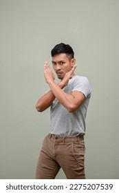 Serious millennial Asian man in casual clothes with crossing hands gesture stands against a green studio background. prohibit, saying no, stop, forbid, ban, disallow, inhibit, dislike, disagree. - Shutterstock ID 2275207539