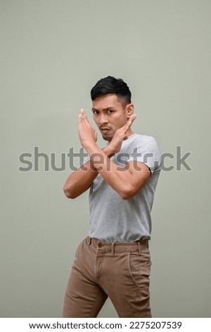 Serious millennial Asian man in casual clothes with crossing hands gesture stands against a green studio background. prohibit, saying no, stop, forbid, ban, disallow, inhibit, dislike, disagree.