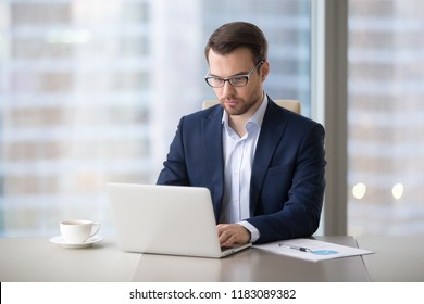 Serious Millennial Businessman Working At Laptop Typing Business Letter Drinking Morning Coffee In Modern Office, Focused Male Manager Busy Checking Email Or Browsing Internet At Computer