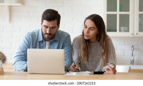 Serious Millennial Couple Planning Family Budget Use E-bank App On Laptop, Do Expenses Calculation, Make Loan Payment Through Online Service, Expenditures Analysis, Writes Notes Sit In Kitchen At Home