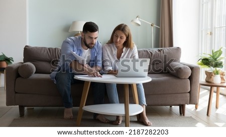 Serious millennial couple sit on sofa sorting out papers, reviewing bills for utility expenses, analysing taxes, do household payment use e-bank application on laptop. Manage finances concept
