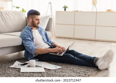 Serious Millennial Jewish Man Using Laptop Sitting On Floor Carpet Leaning On Couch, Focused Casual Guy Communicating Online, Writing Emails, Distantly Working Or Studying On Computer Doing Paperwork