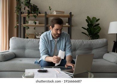 Serious millennial male in glasses focused on paying utility bills taxes rental charges online using laptop. Young man working from home office sit on couch hold paper invoice check information online