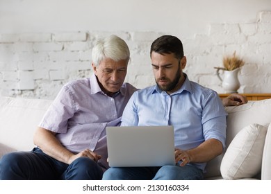 Serious Millennial Man Explain, Help To His Older 60s Dad With Laptop Usage, Sit Together On Sofa At Home Look At Wireless Computer Showing New Software Buy On E-commerce Services. Modern Tech Concept