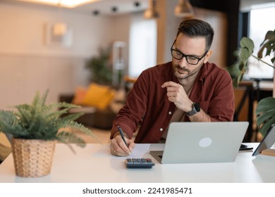 Serious millennial man using laptop sitting at the table in a home office. Focused guy in casual clothing looking at the paper, writing down ideas or a future business plan and using laptop. - Powered by Shutterstock