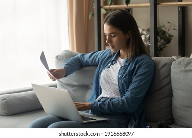 Serious millennial woman sit on couch hold laptop work with electronic document and paper hardcopy of loan insurance contract. Focused young female read official letter check actual information online