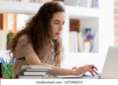 Serious millennial woman using computer sitting at classroom table, focused girl e-learning using online tasks preparing for test exams, typing emails, distantly working or studying on laptop indoors