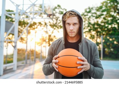 Serious Minded Sunlit Young Sporty Sportsman Man 20s Wear Grey Sports Clothes Hood Look Camera Training Holding In Hand Ball Play At Basketball Game Playground Court. Outdoor Courtyard Sport Concept.