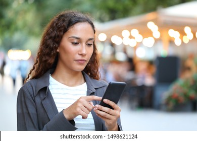 Serious mixed race woman using smart phone walking in the street with city lights in the background