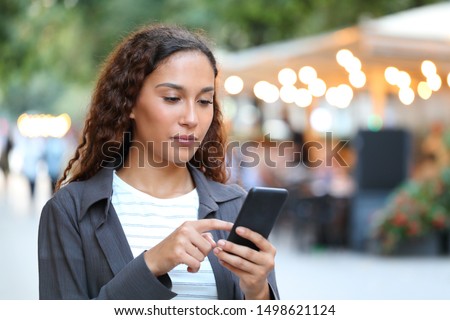 Serious mixed race woman using smart phone walking in the street with city lights in the background