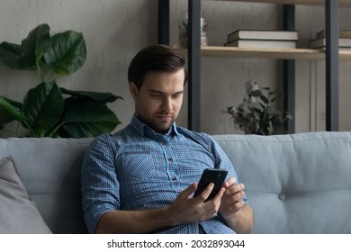 Serious mobile phone user reading text message on screen, resting on couch et home. Millennial man focused on smartphone, using online app, virtual service for payment, shopping, chatting on internet