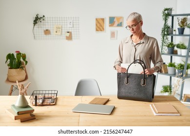 Serious modern middle-aged businesswoman in glasses standing at table and putting folder into bag while going to meeting
