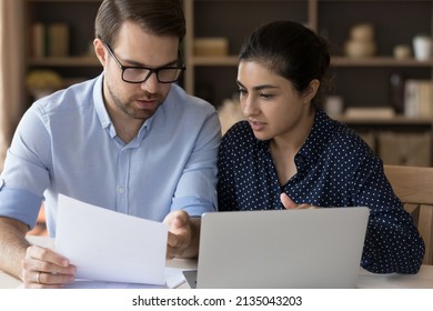Serious multiethnic business partners coworkers students Indian woman Caucasian man sit at desk discuss document read sales report. Two young diverse teammates engaged in paperwork solve problem talk