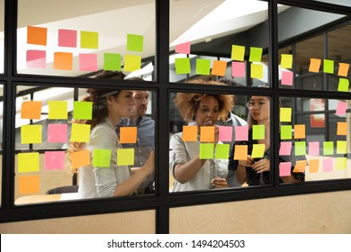 Serious multiracial business team people brainstorm on corporate project, african female leader coach explain creative idea work plan write on post in sticky notes stand behind glass office wall - Powered by Shutterstock