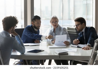 Serious multiracial older and young businesspeople gathered in boardroom discuss financial statistics, analyze sales report, forecasting work together at office meeting. Teamwork, negotiations concept