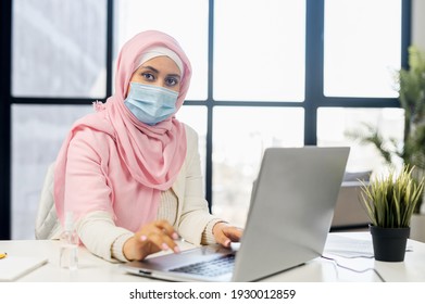 Serious Muslim Woman Wearing Hijab And Protective Face Mask Using Laptop In Modern Office Space. Islamic Businesswoman In Surgical Mask Looks At The Camera Sitting At The Desk