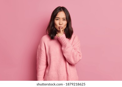 Serious mysterious brunette Asian woman presses index finger to lips makes hush gesture tells secret asks to be quiet wears long sleeved jumper poses against pink background. Secrecy concept