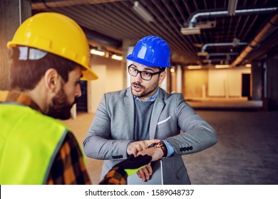 Serious Nervous Supervisor Arguing With His Worker And Pointing At Wristwatch. Work Must Be Done On Time With No Excuses. Building In Construction Process Interior.