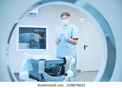 Serious Nurse In Protective Mask And Gloves Using Tablet And Reading Patients Card While Preparing MRI Scanner For Covid Patient