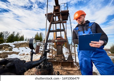 Serious Oil Man In Work Overalls And Helmet Holding Clipboard And Looking At Oil Pump Rocker-machine. Male Worker Checking Oil Pumping Unit, Making Notes. Concept Of Oil Extraction, Petroleum Industry