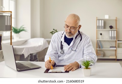 Serious Old Caucasian Doctor In Medical Uniform And Glasses Work On Computer In Hospital Write Prescription. Mature Male GP Or Therapist Use Laptop Prescribe Meds To Patient In Card Or Journal.