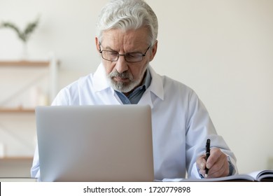 Serious Old Doctor Therapist Making Notes In Medical Journal Doing Online Research Using Laptop In Hospital Office. Senior Adult Male Physician Working On Computer Writing Prescription Sits At Desk.