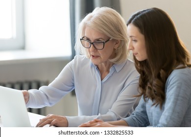 Serious Old Female Mentor Teacher Coach Teaching Intern Or Student Computer Work Pointing At Laptop, Mature Executive Manager Explaining Online Project To Young Employee Learning New Skills In Office