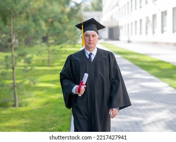 Serious Old Man In Graduation Gown Holding Diploma Outdoors.