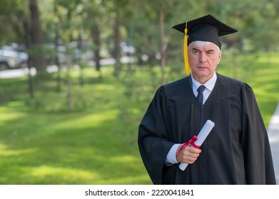 Serious Old Man In Graduation Gown Holding Diploma Outdoors.