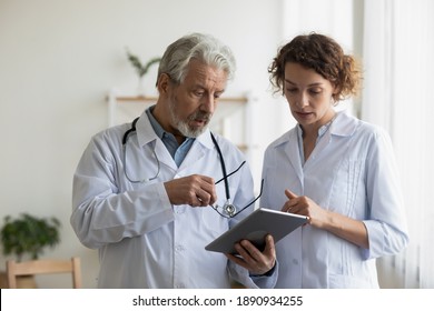 Serious Old Mature Male Doctor Giving Instructions To Focused Young Colleague, Discussing Illnesses Treatment Or Health Test Results On Digital Computer Tablet In Office, Medical Cooperation Concept.