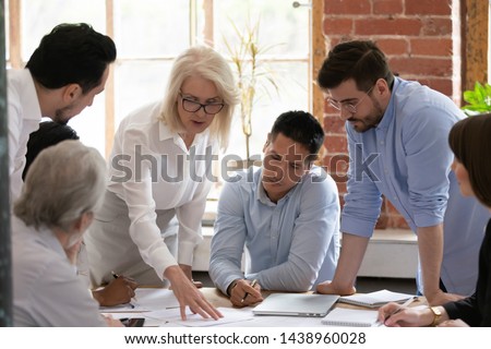 Serious old mature woman team leader coach teach young workers explain paper business plan at group meeting, focused senior female teacher mentor training diverse staff at corporate office workshop