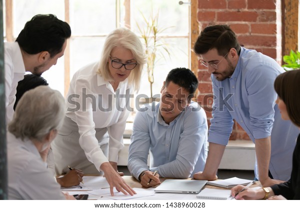 Serious old mature woman team leader coach teach\
young workers explain paper business plan at group meeting, focused\
senior female teacher mentor training diverse staff at corporate\
office workshop
