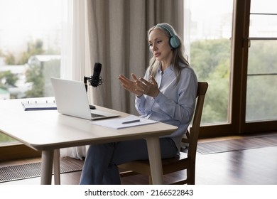 Serious older 50s woman in wireless headphones sit at desk with laptop make speech into mike, holding on-line class, teach internet audience, share skill experience during livestream. Webinar concept