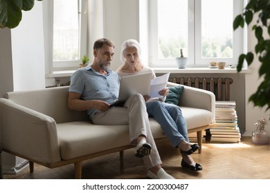 Serious Older 60s Couple Paying Paper Bills On Internet, Using Online Banking App On Laptop, Checking Documents Together, Counting Loan, Insurance, Tax Fees, Planning Budget, Discussing Expenses