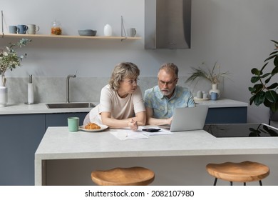 Serious Older 60s Married Couple Discussing Budget, Income, Expenses, Calculating Money, Counting Insurance Fees At Calculator, Doing Paperwork Together. Paying Bills, Taxes, Using Laptop In Kitchen