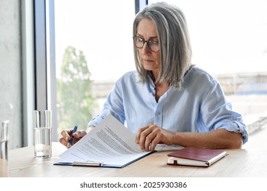 Serious older lawyer businesswoman in glasses reading signing trust partnership contract sit at table in office. Executive ceo put signature make legal bank sale financial loan investment agreement.