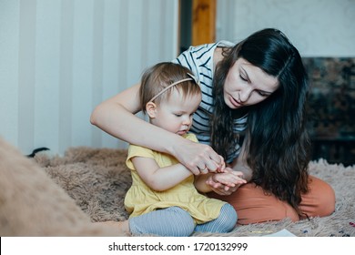 Serious One Year Old Girl Sitting On The Bed And A Concentrated Mother Holding Her Hands