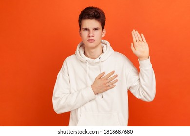 Serious Patriotic Male In White Sweatshirt With Hood Holding Hand On Heart, Juryman Swearing To Speak Truth In Court, Honor And Conscience. Indoor Studio Shot Isolated On Orange Background