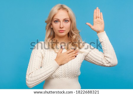 Serious patriotic woman with curly blond hair raising one arm and putting on chest another making oath, swearing. Indoor studio shot isolated on blue background