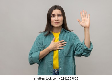 Serious Patriotic Woman Holding Hand On Heart, Juryman Swearing To Speak Truth In Court, Honor And Conscience, Wearing Casual Style Jacket. Indoor Studio Shot Isolated On Gray Background.